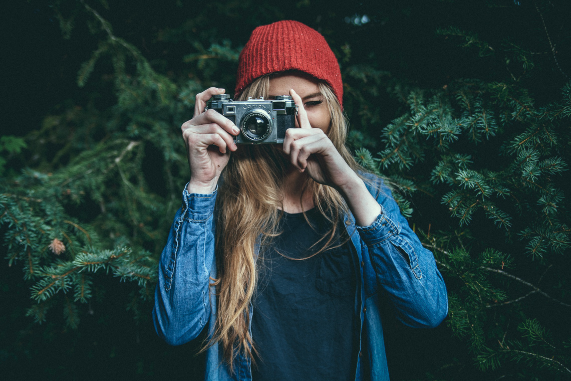 Woman holding a film camera up to her eye. She is standing in a fir tree.