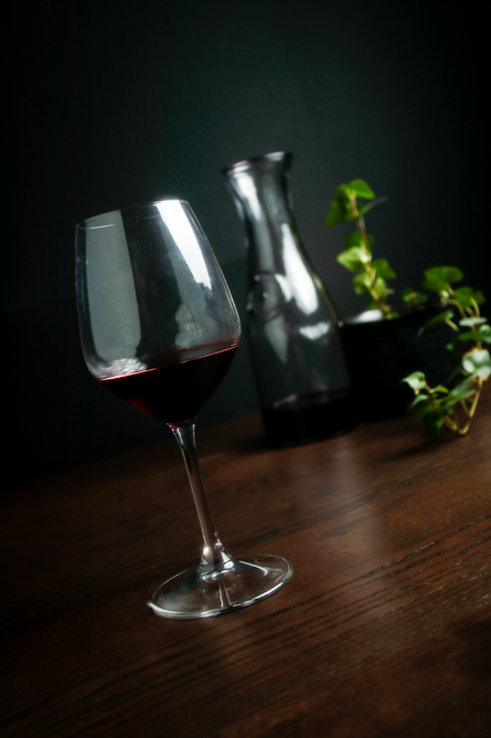 A glass of red wine on a wooden table with a pitcher and plant in the background.
