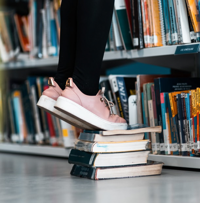 Person in pink shoes standing on books to reach a shelf