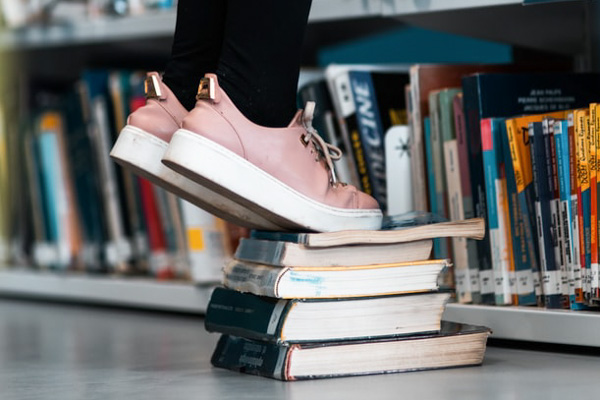 Person in pink shoes standing on books to reach a shelf