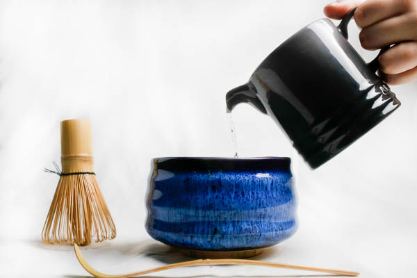 A hand pouring water from a grey pitcher into a matcha bowl