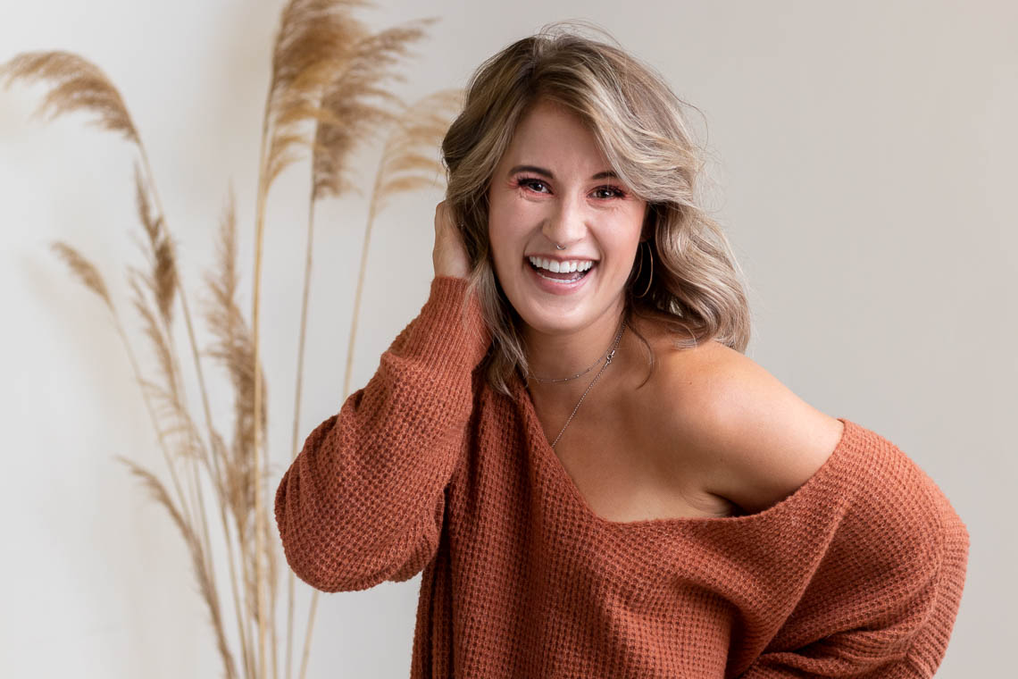 Blonde woman in an orange sweater laughing with her hand in her hair.