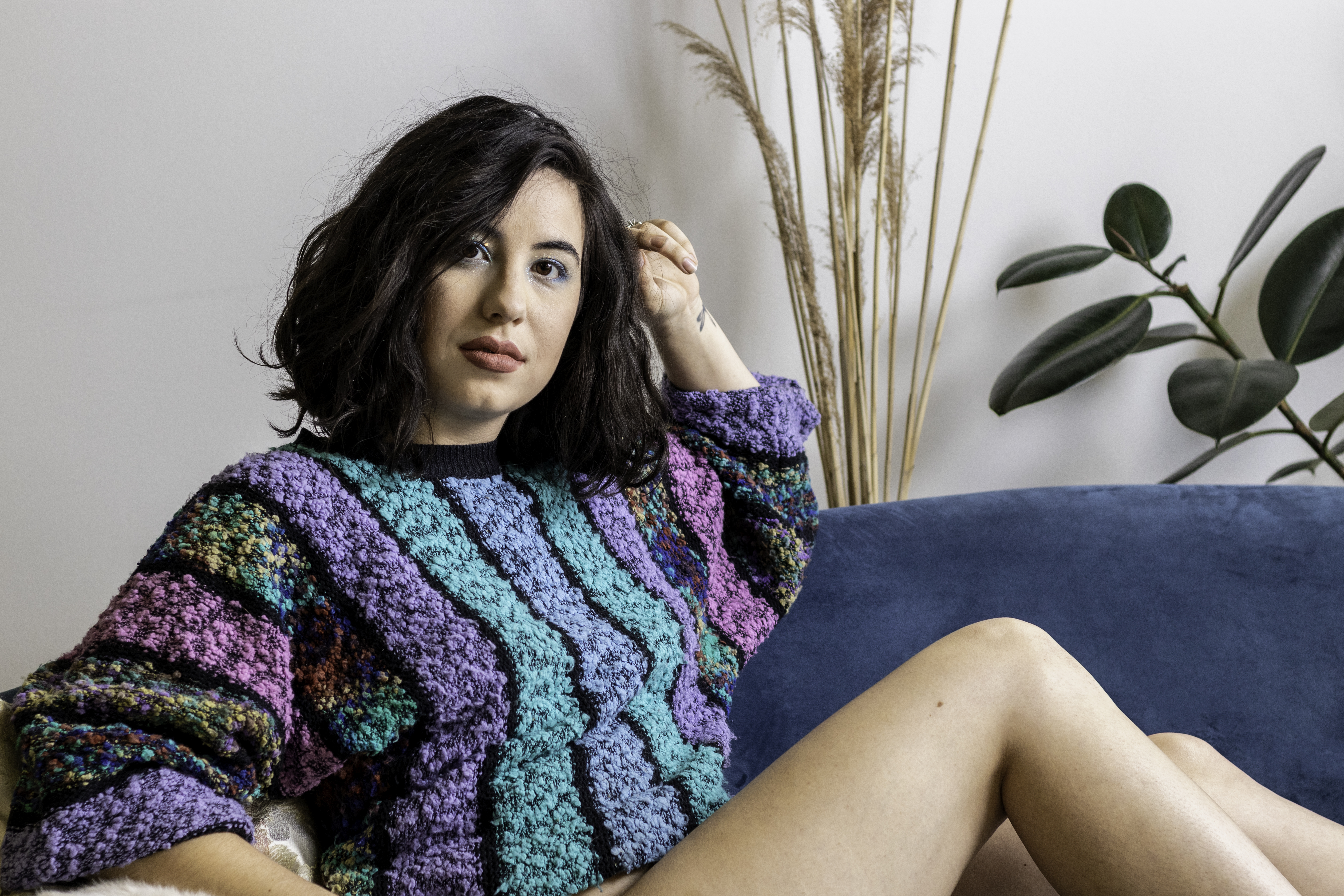Black-haired woman in a colourful sweater looking at the camera and lounging on a blue velvet couch.