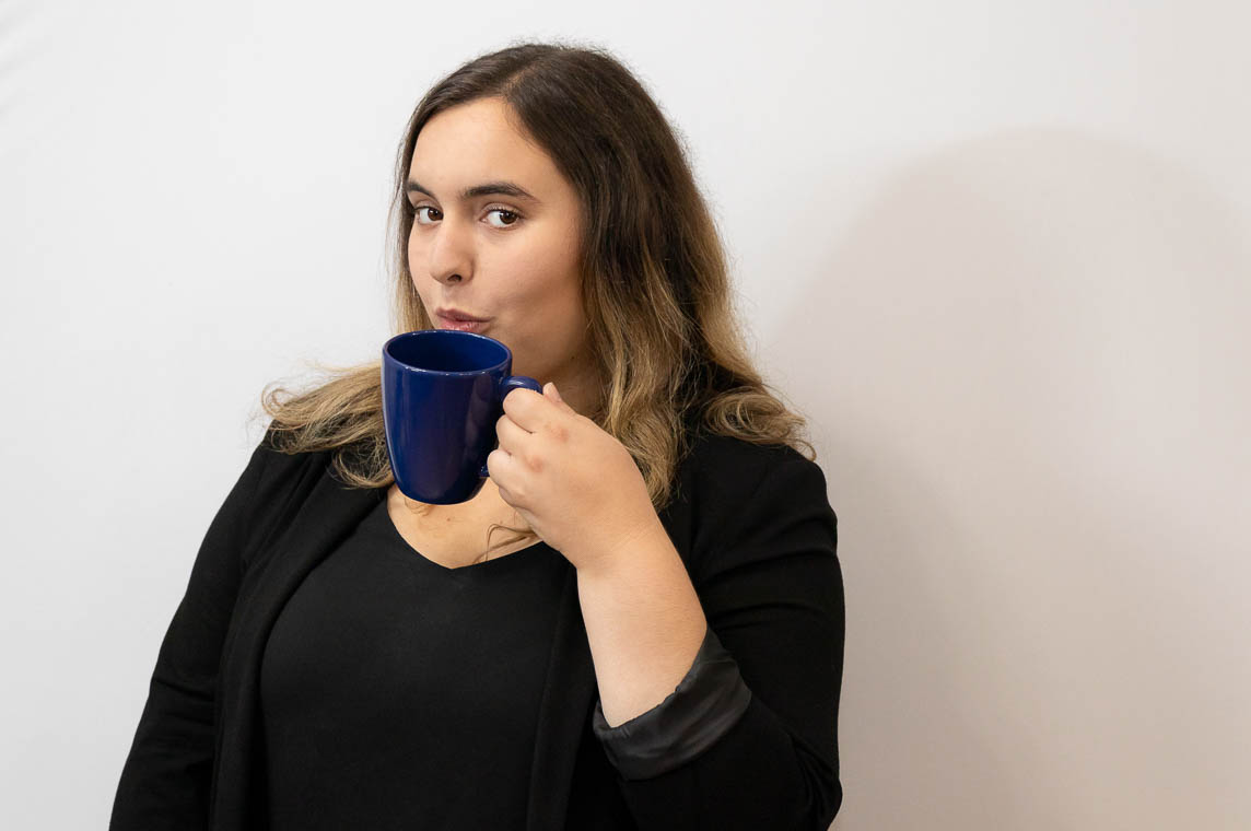 Brunette woman wearing black in front of a white backgrond. She is holding a coffee mug.