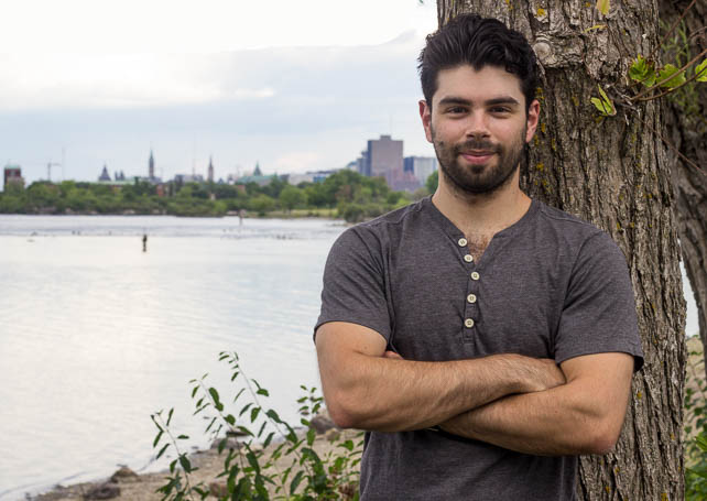 Man with black hair and a beard crossing his arms. He is outside and looking at the camera.