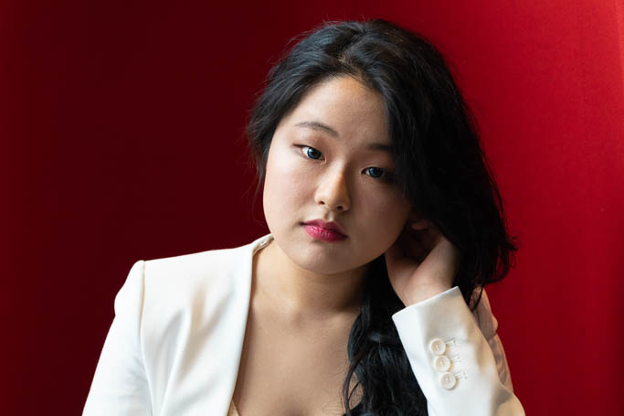 Asian woman wearing a white blazer and looking at the camera. She is in front of a red background.