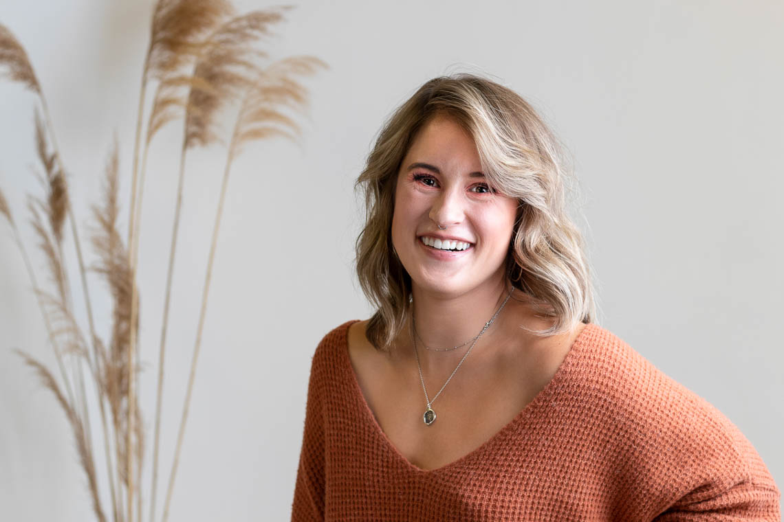 Blonde woman looking at the camera and smiling. She is wearing an orange sweater and is in front of a grey background.