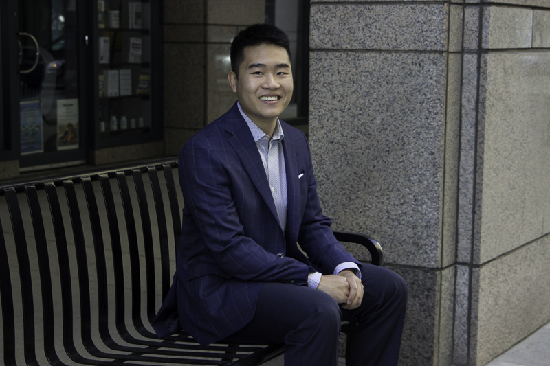 An Asian man in a navy suit sitting on a bench and smiling at the camera.
