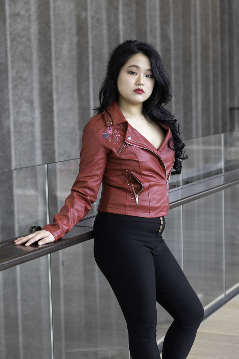 An Asian woman modeling with a grey background wearing black pants and a red leather jacket.