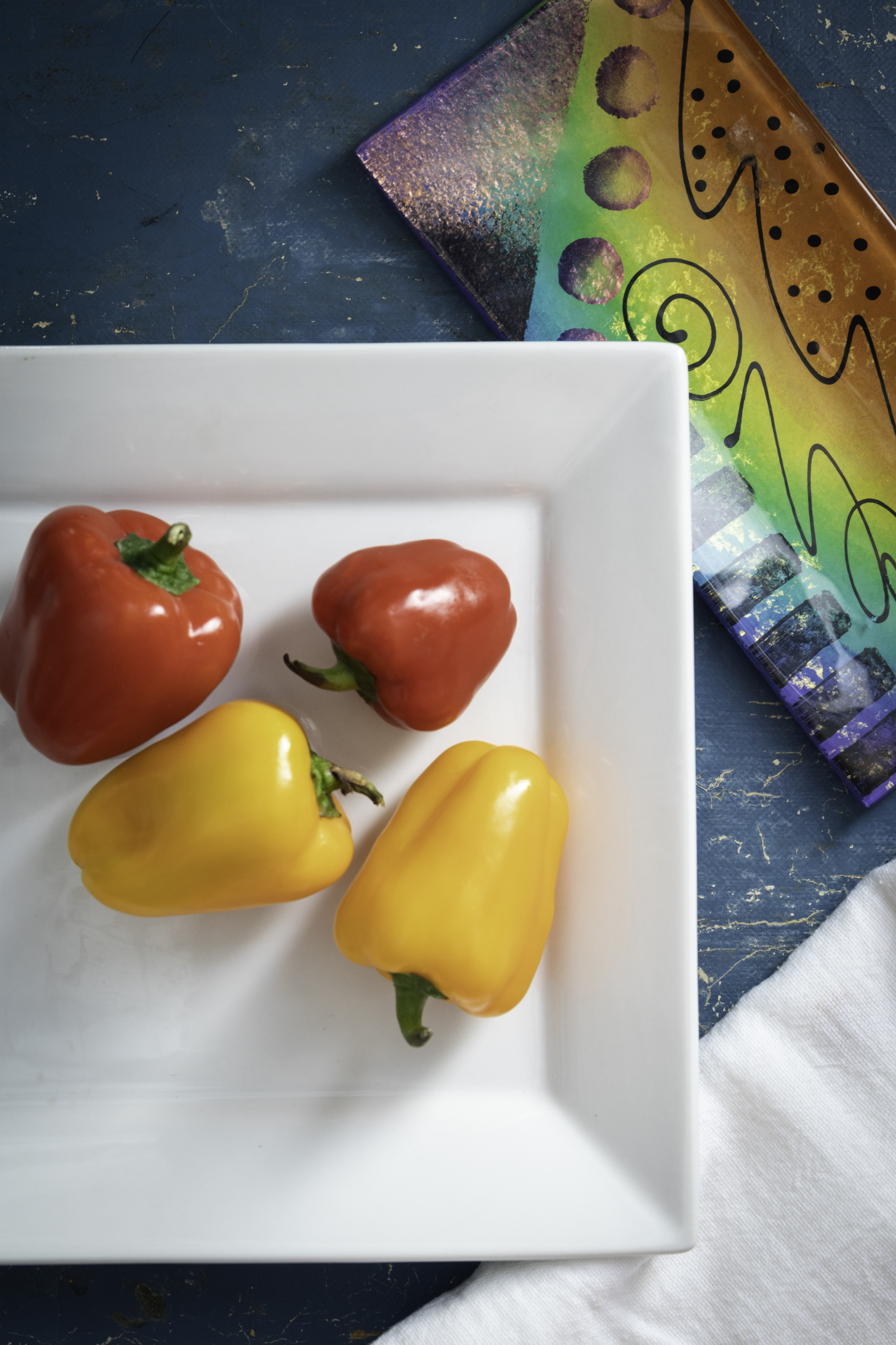 Red and yellow peppers on a white plate with a blue background and a colourful plate in the background.