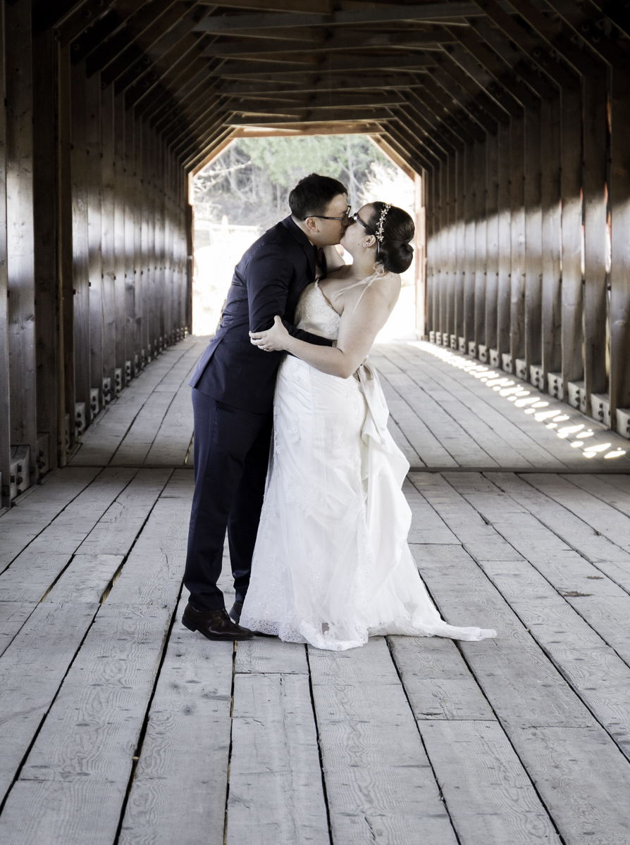 Man and woman in wedding attire kissing on a covered bridge.