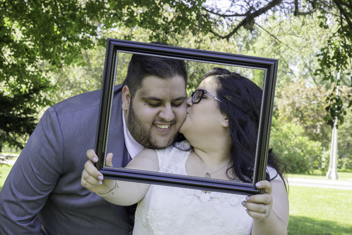 A bride holding a frame around her and her groom's faces as she kisses him on the cheek.