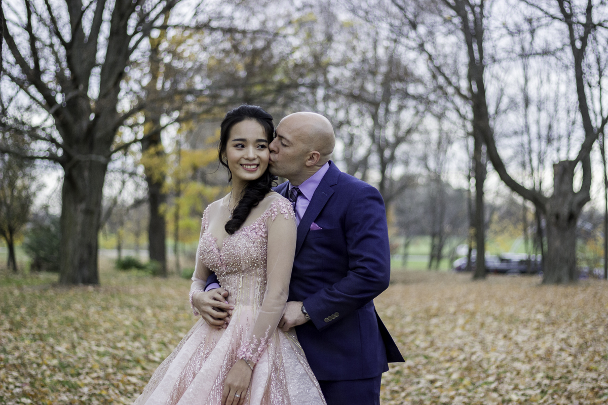 Man in a blue suit kissing bride in a pink dress with an autumn background.