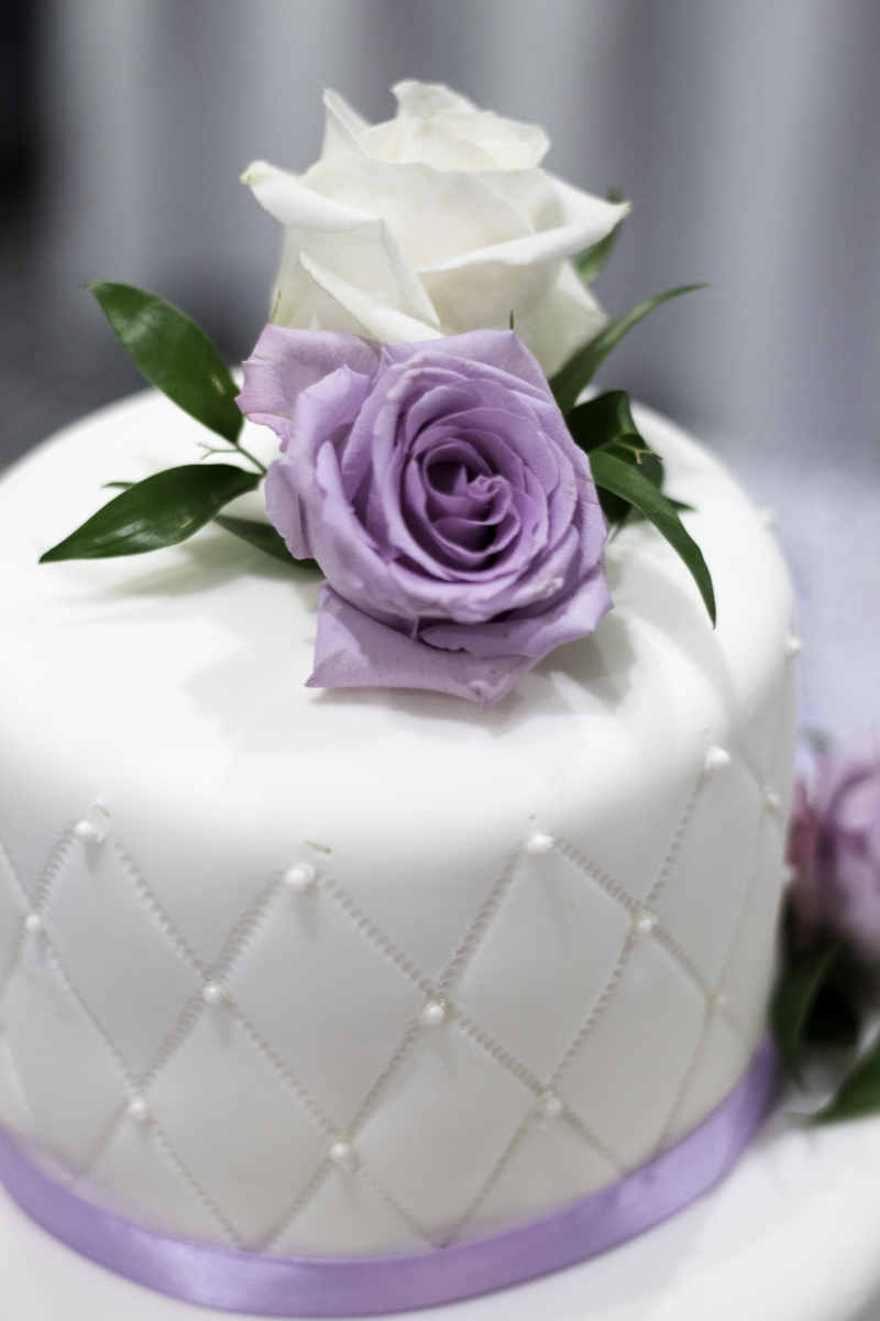 Close up of a white and lilac wedding cake with roses on top.